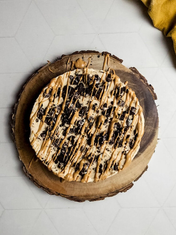 A round dessert topped with crushed cookies and drizzled caramel, placed on a wooden board.