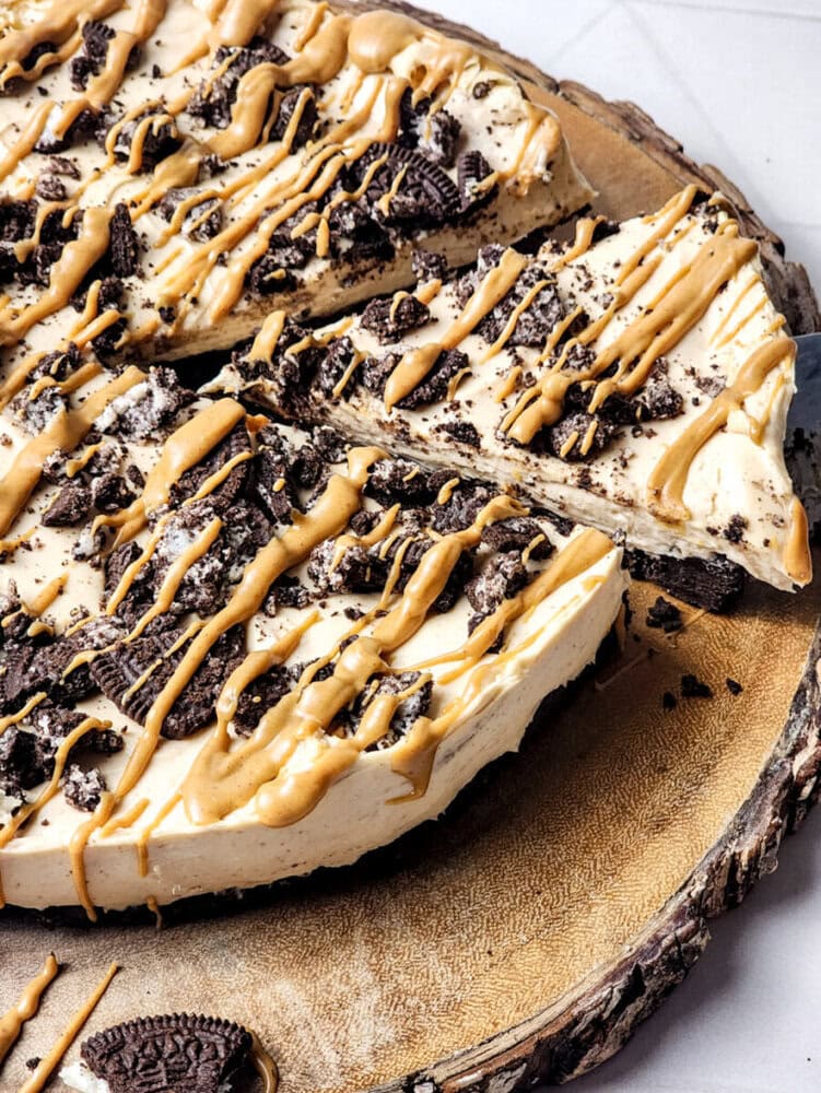 A peanut butter cheesecake topped with chocolate cookie pieces and drizzled with peanut butter, sliced and displayed on a wooden platter.