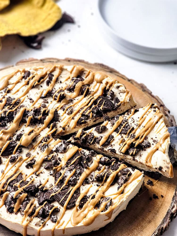 A cheesecake topped with crushed cookies and caramel drizzle is sliced, set on a wooden board with plates in the background.