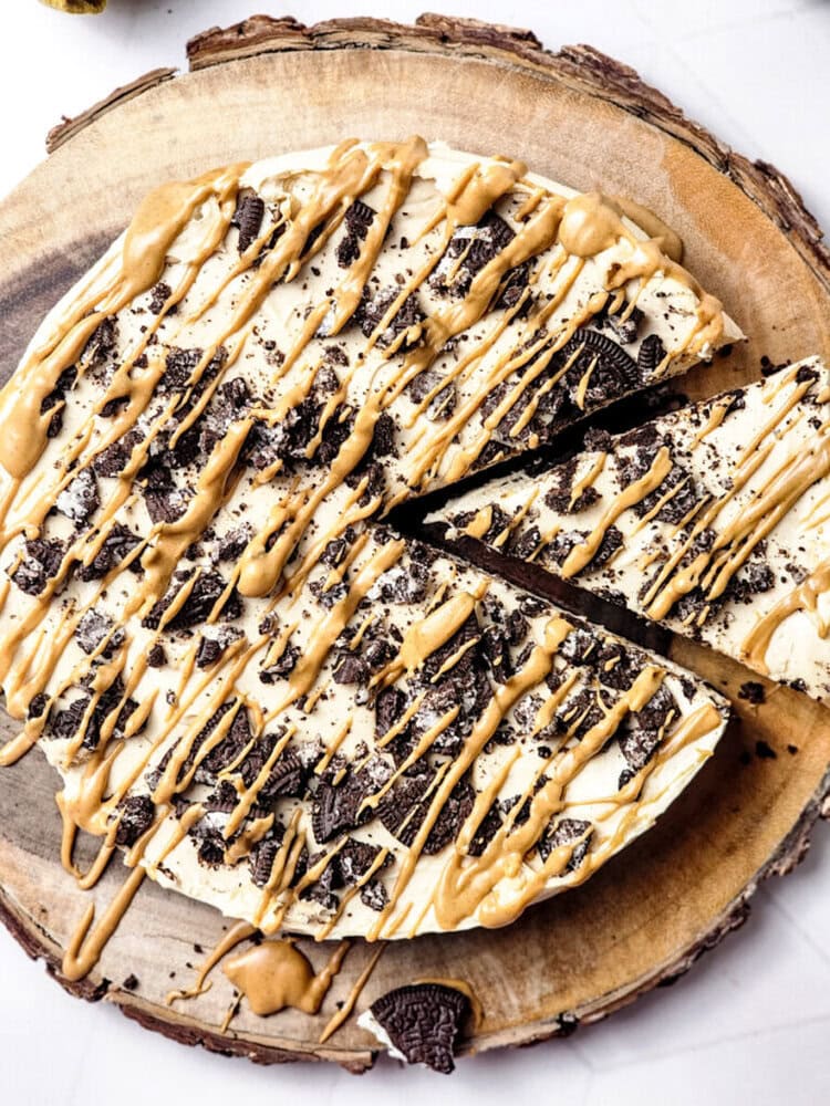 A round dessert pizza topped with crushed cookies and drizzled with peanut butter sauce, placed on a wooden slab. One slice is partially separated.
