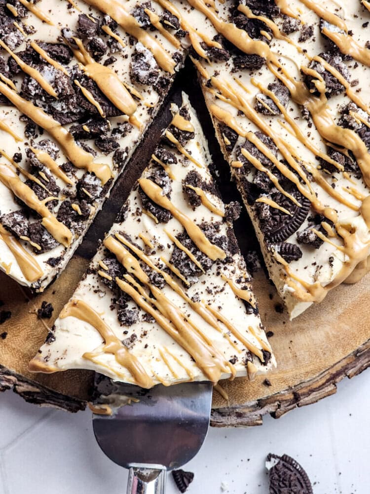 A slice of cheesecake topped with crushed cookies and peanut butter drizzle is being lifted with a spatula. The dessert rests on a wooden board.