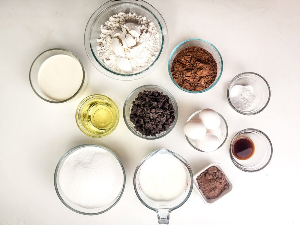 Top-down view of baking ingredients including flour, cocoa powder, sugar, chocolate chips, eggs, oil, milk, vanilla extract, and baking powder arranged on a white surface.