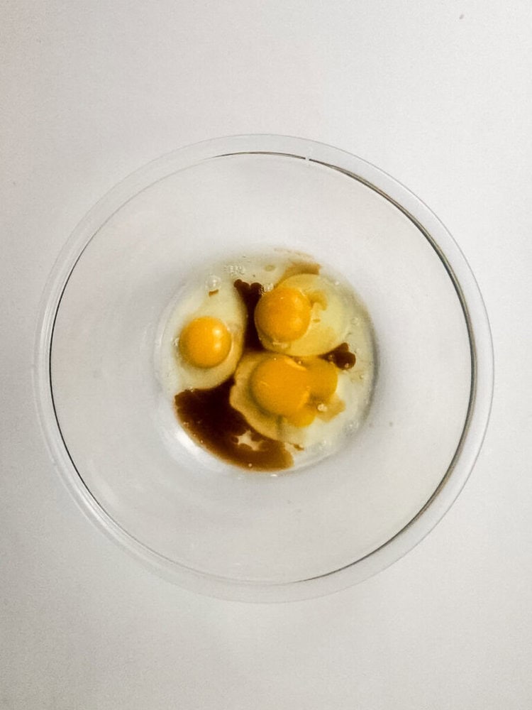 A clear bowl containing three raw eggs, vanilla extract, and sugar on a white surface, viewed from above.