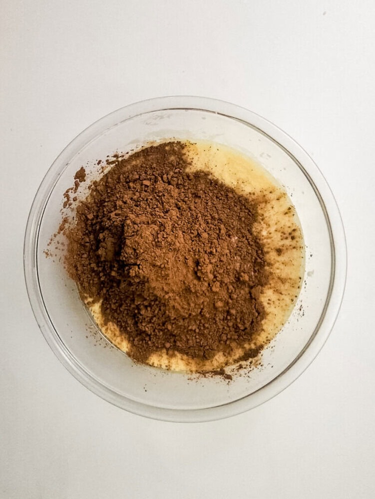 A clear bowl containing a yellow batter with cocoa powder sprinkled on top, placed on a white surface.