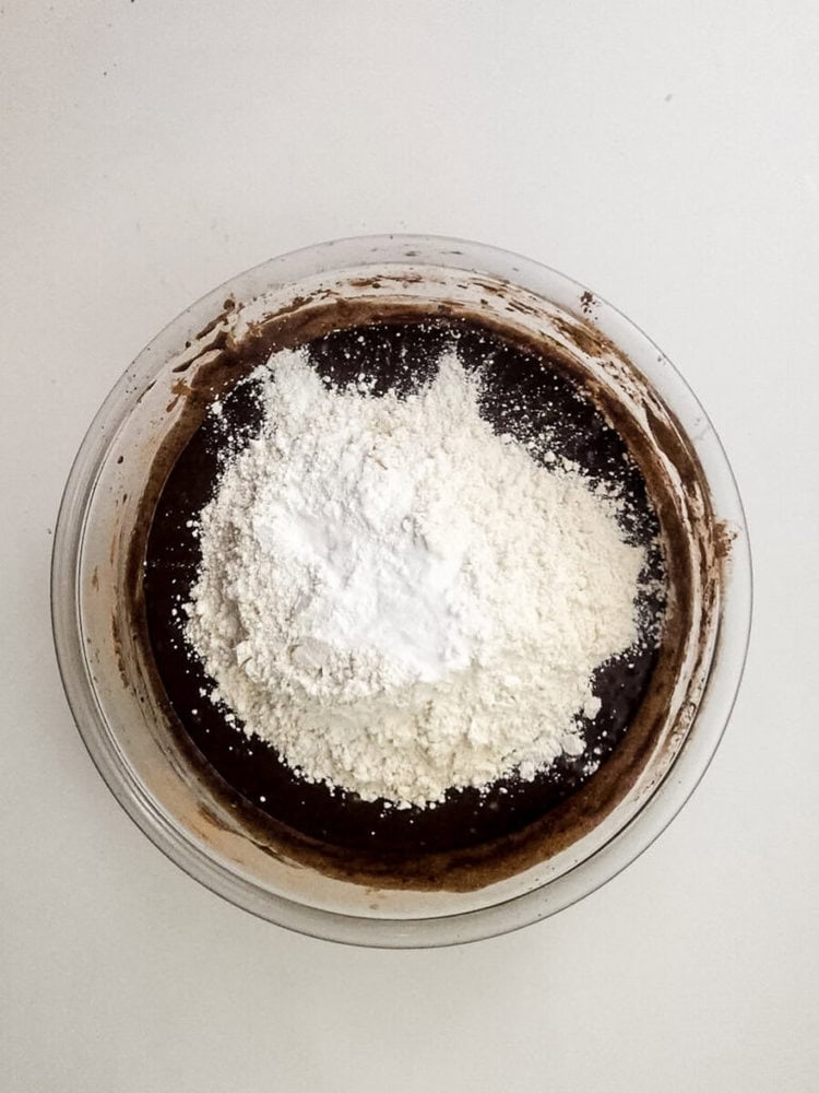 A glass bowl containing chocolate batter topped with a pile of flour and baking powder on a white surface.