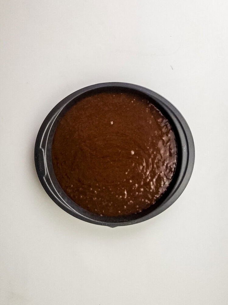 A round black baking pan filled with unbaked chocolate cake batter sits on a plain white surface.