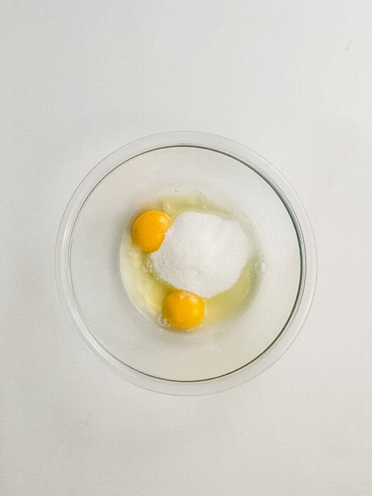A clear glass bowl containing two raw eggs and a mound of white sugar on a white surface.