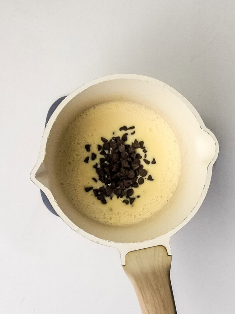 A white saucepan contains a creamy yellow mixture with a pile of chocolate chips in the center, viewed from above on a white background.