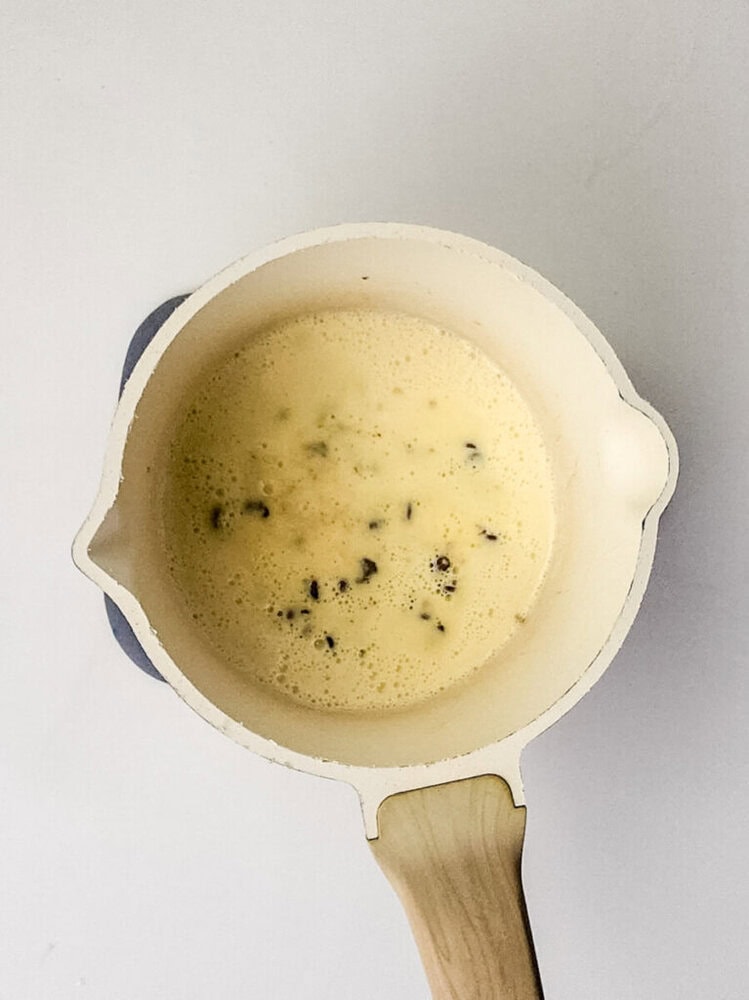 A cream-colored saucepan with a light wooden handle contains a pale yellow, foamy mixture with small dark specks on a white background.