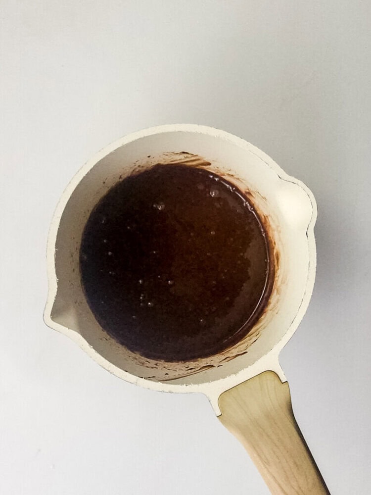 A white saucepan with a wooden handle contains a dark brown liquid mixture, possibly melted chocolate or sauce, viewed from above on a white surface.