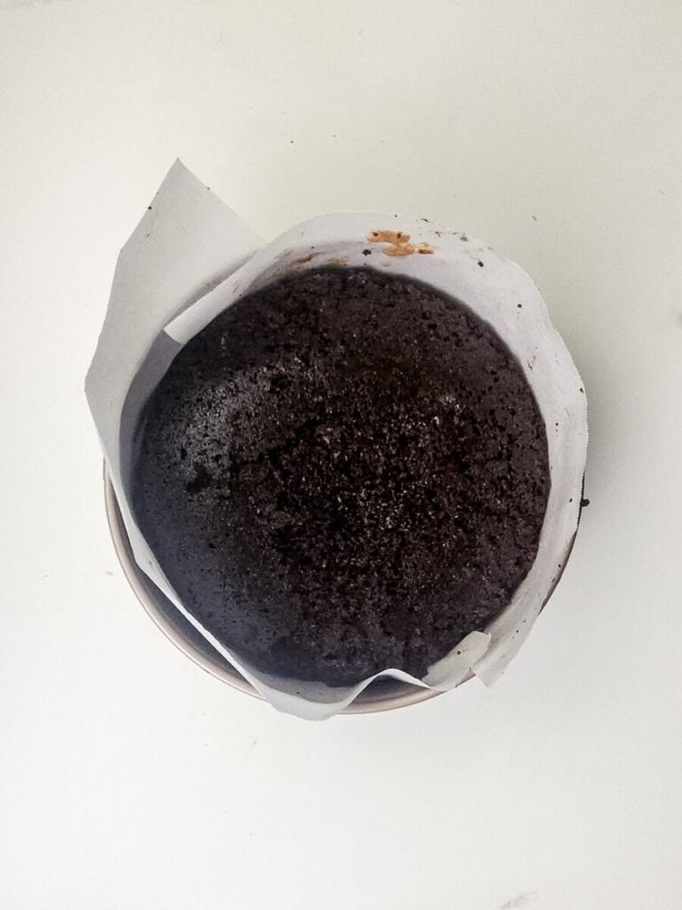 A round chocolate cake with a dark, textured top sits in a bowl lined with white parchment paper on a white surface.