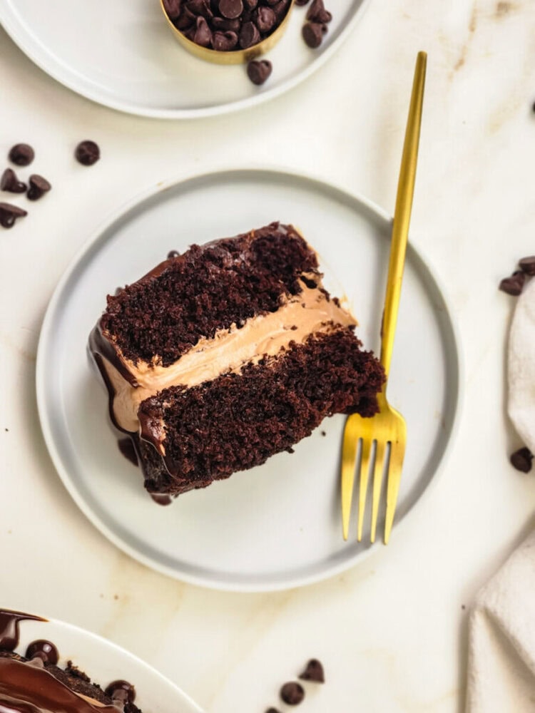 A slice of chocolate layer cake with chocolate frosting on a white plate, accompanied by a gold fork, with chocolate chips scattered nearby.