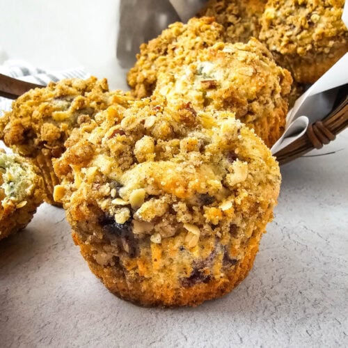 Close-up of a basket with several crumb-topped muffins, featuring a visible muffin in the foreground on a light surface.