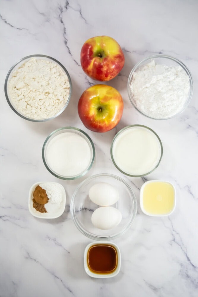 Ingredients laid out on a marble surface include two apples, flour, sugar, milk, eggs, oil, vanilla extract, baking powder, and cinnamon.