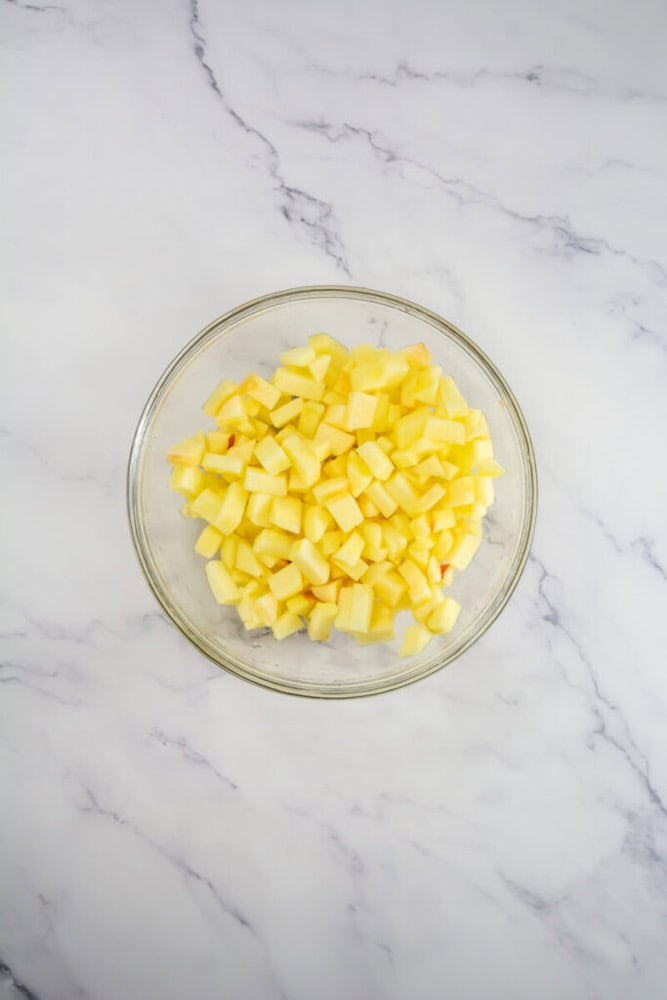 Glass bowl filled with diced yellow fruit, possibly apples, on a marble surface.
