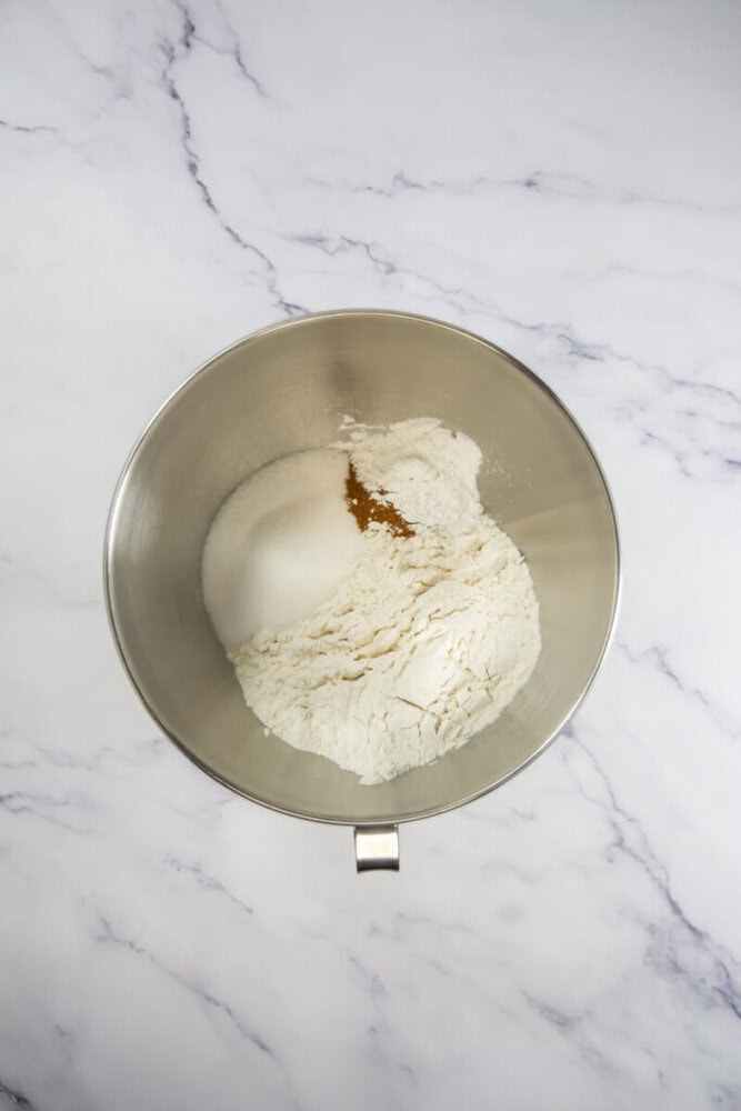A stainless steel mixing bowl containing flour and brown sugar on a marble countertop.