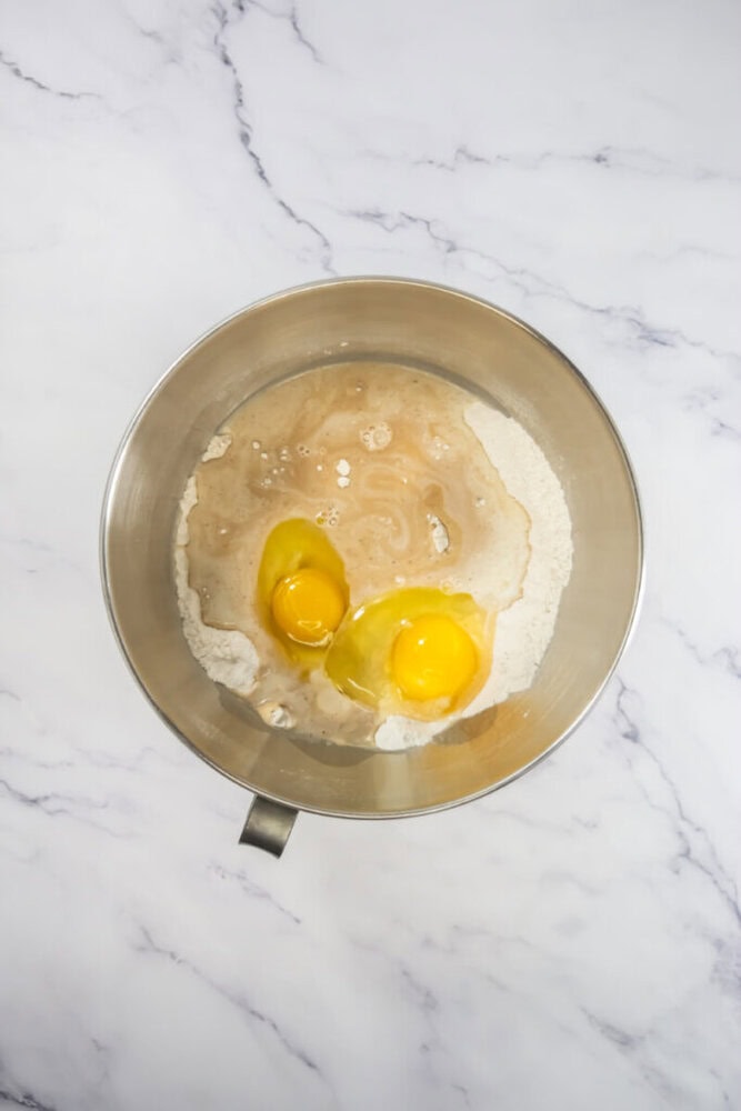 A mixing bowl containing flour, two cracked eggs, and liquid ingredients on a marble surface.