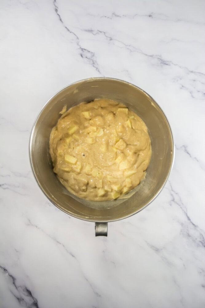 Apple cake batter with apple chunks in a metal mixing bowl, placed on a white marble surface.