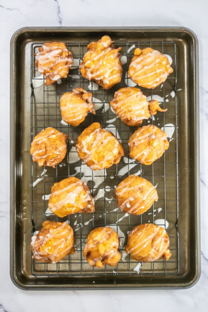 Twelve glazed apple fritters on a wire rack atop a baking sheet, with some icing drips on the sheet.