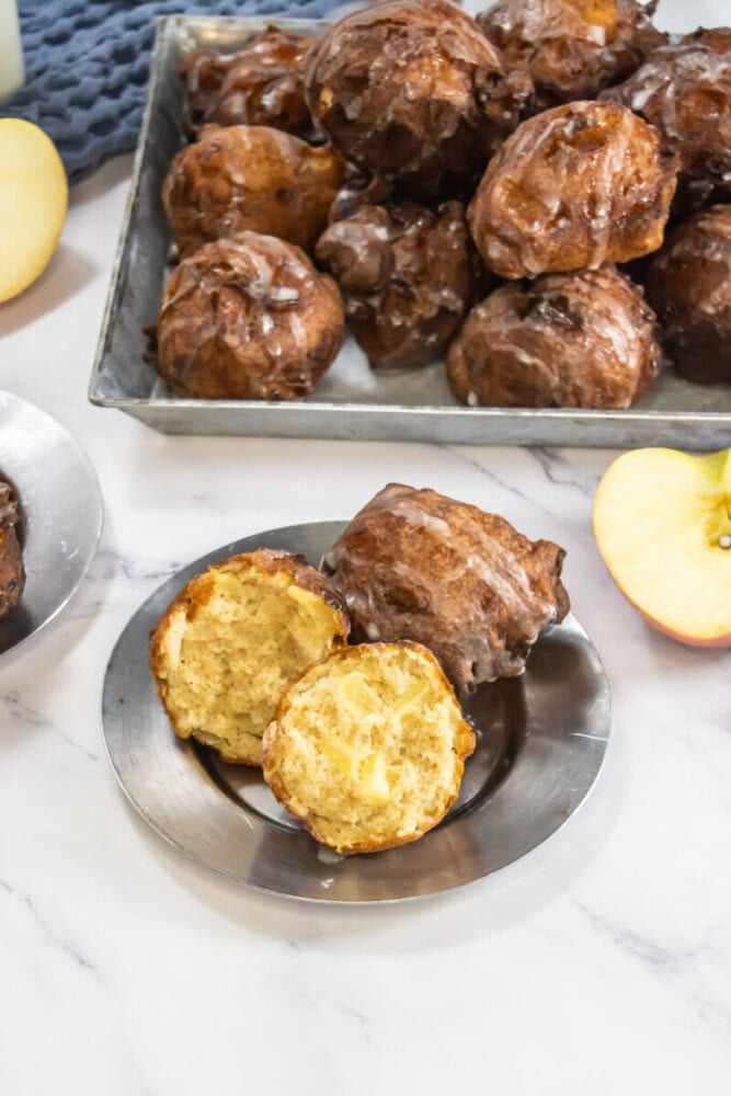 A tray of glazed apple fritters is in the background. In the foreground, a metal plate holds a split apple fritter, showcasing its soft interior. Slices of apple are beside the plate.