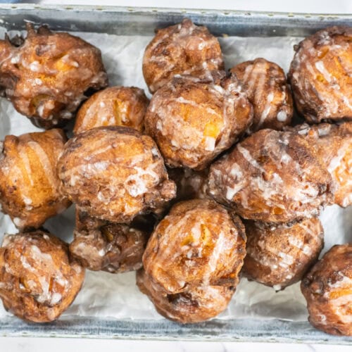 A metal tray filled with glazed apple fritters on a white paper liner.