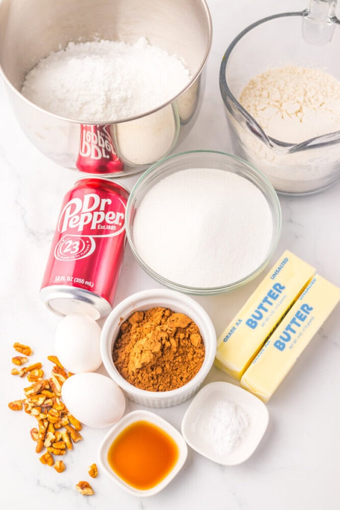 An assortment of baking ingredients including flour, sugar, cocoa powder, eggs, vanilla, butter, baking soda, pretzels, and a can of Dr Pepper arranged on a countertop.