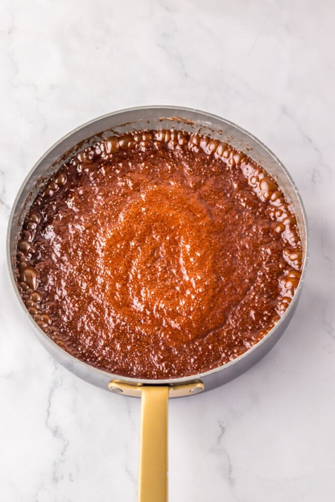A saucepan filled with bubbling brown sauce or caramel sits on a white marble surface.