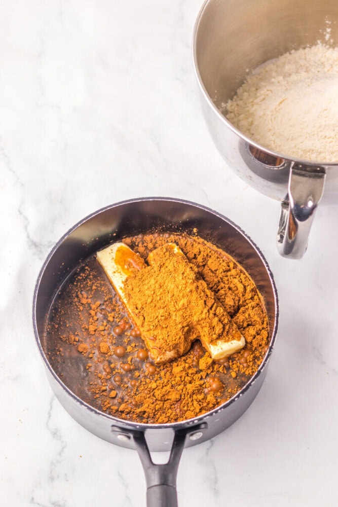 A saucepan containing butter, cocoa powder, and liquid sits next to a mixing bowl of flour on a marble surface.