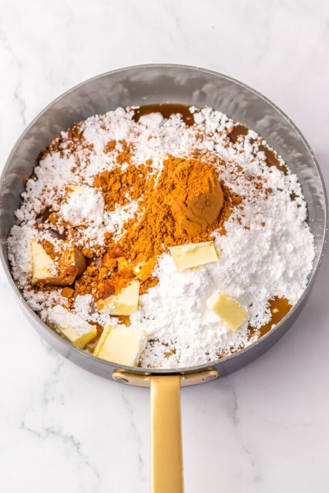 A saucepan filled with powdered sugar, cocoa powder, and several pieces of butter on a marble surface.