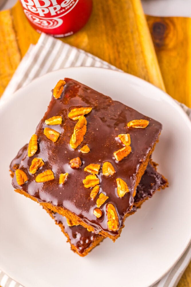 Two square pieces of chocolate-glazed cake topped with pecans are stacked on a white plate, with part of a Dr Pepper can visible in the background.