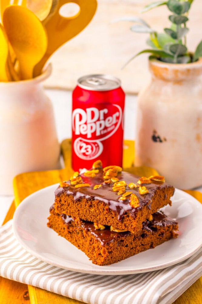 Two pieces of chocolate cake with glaze and nuts are stacked on a plate, with a can of Dr Pepper and kitchen utensils in the background.