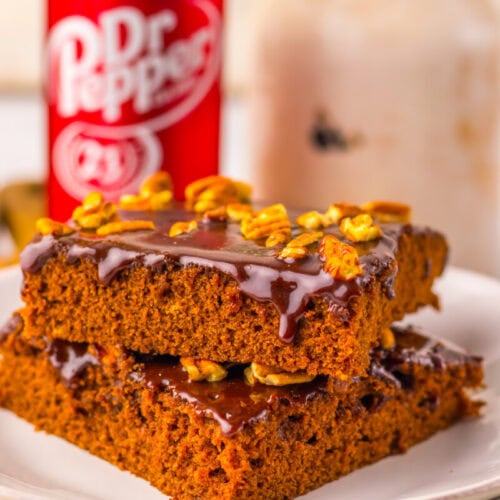 Two pieces of chocolate-drizzled cake topped with pecans are stacked on a plate, with a can of Dr Pepper and a glass jar in the background.