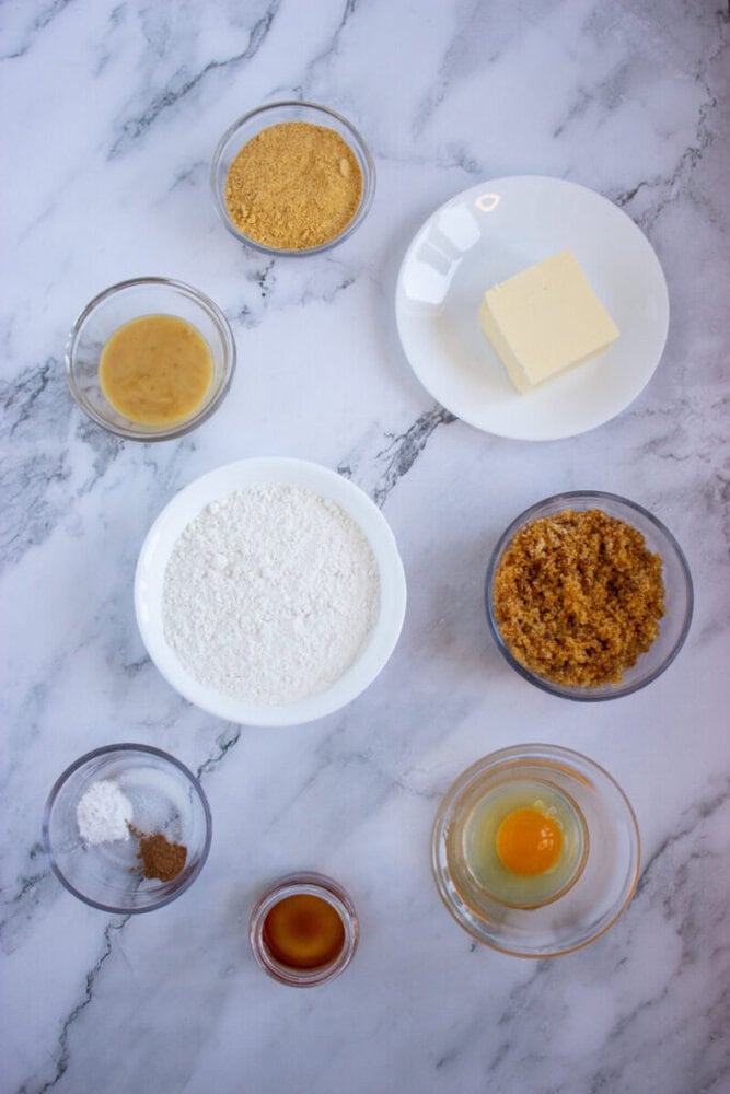 Baking ingredients on a marble surface: butter on a plate, bowls of flour, brown sugar, an egg, honey, vanilla extract, baking powder, and spices.