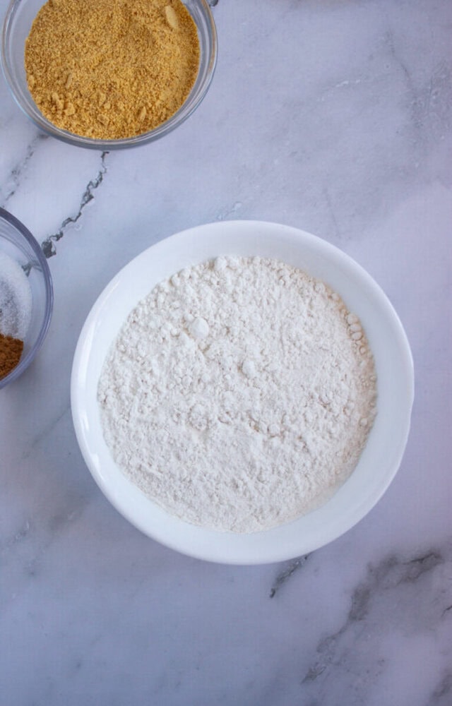 White flour in a white bowl on a marble surface, with additional bowls containing brown powder and sugar partially visible.
