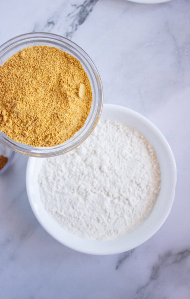 A bowl of white flour sits next to a jar of yellow cornmeal on a marble surface.