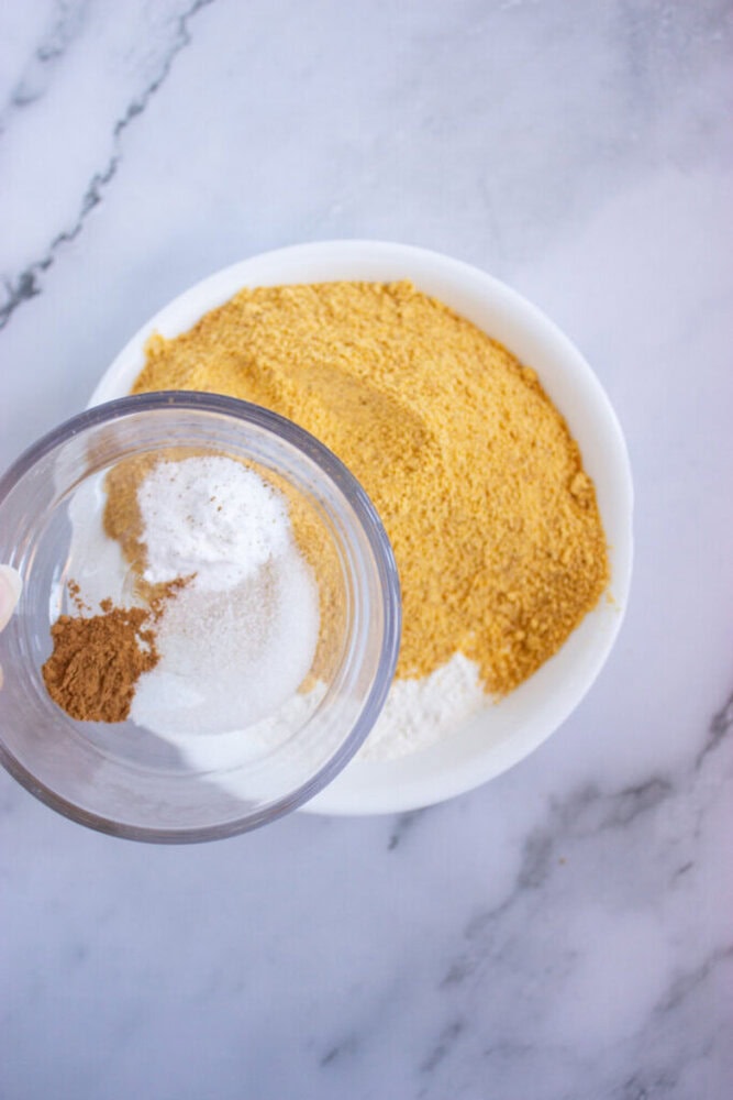 A white bowl filled with yellow cornmeal and white flour. A separate bowl above contains cinnamon, sugar, and baking powder.