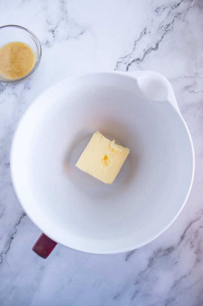 A stick of butter sits in a white mixing bowl on a marble surface, with a small bowl of honey nearby.