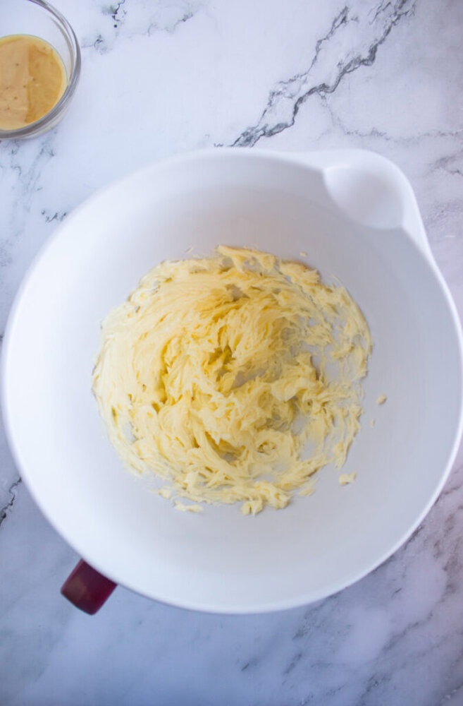 Butter is creamed in a large white mixing bowl placed on a marble countertop. A small bowl with a yellow mixture is in the upper left corner.