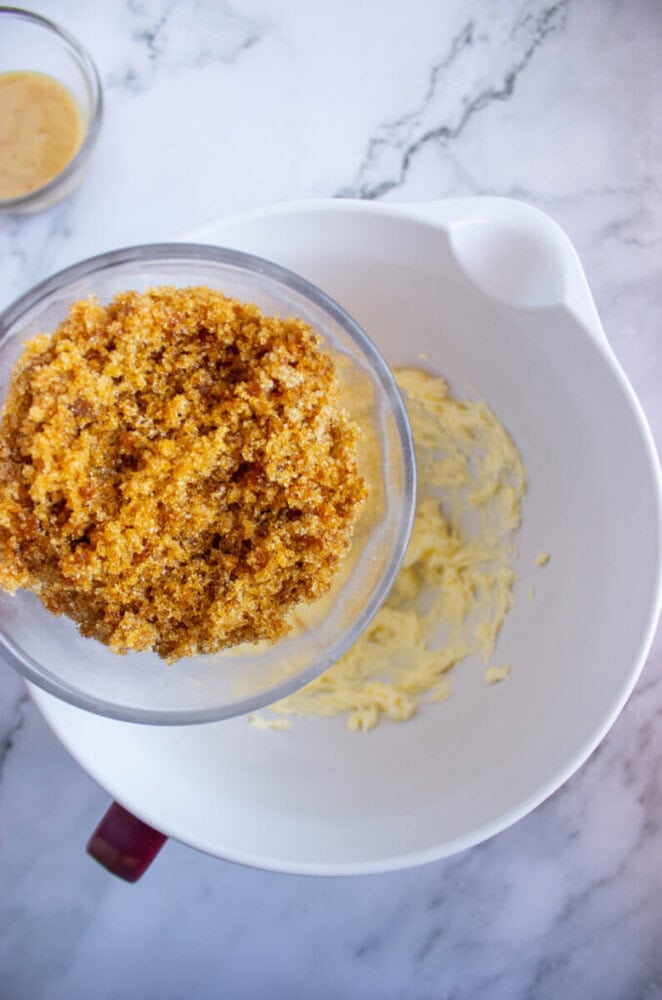 A bowl of creamed butter in a mixing bowl, with a measuring cup of brown sugar about to be added. Marble countertop background.