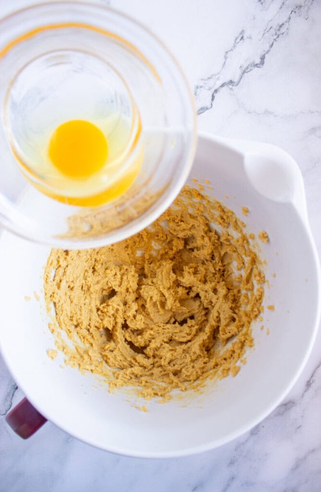 A mixing bowl with dough mixture and an egg in a glass bowl about to be added, on a marble countertop.