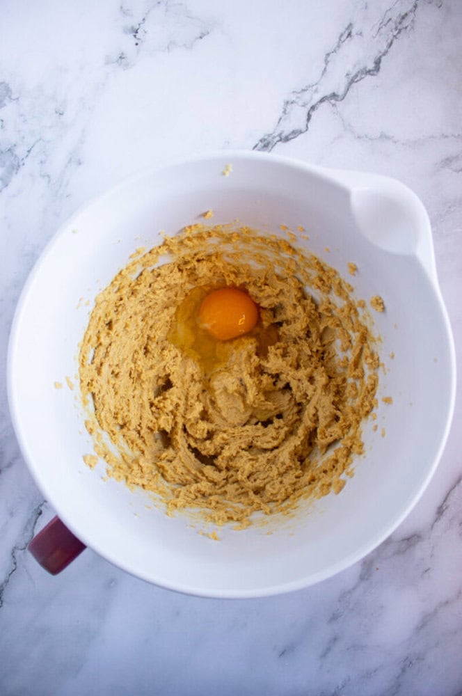 A white mixing bowl with cookie dough mixture and a cracked egg on a marble countertop.