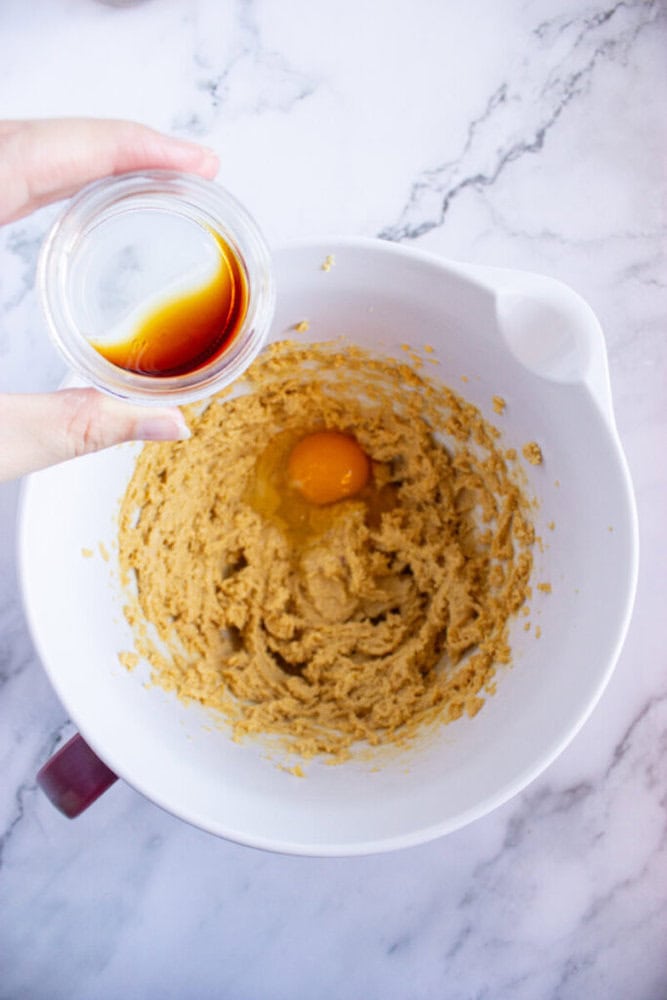 Bowl with cookie dough and a cracked egg, held over by a hand with a small glass bowl of vanilla extract, on a marble surface.