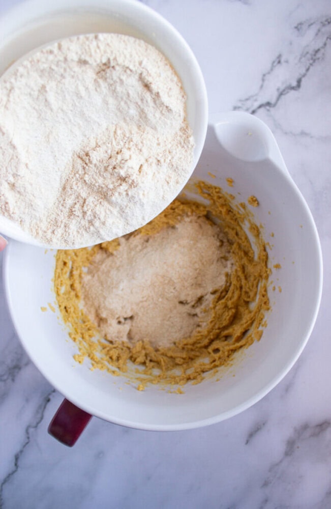 Bowl of flour being added to a large mixing bowl with a creamy batter, placed on a marble surface.