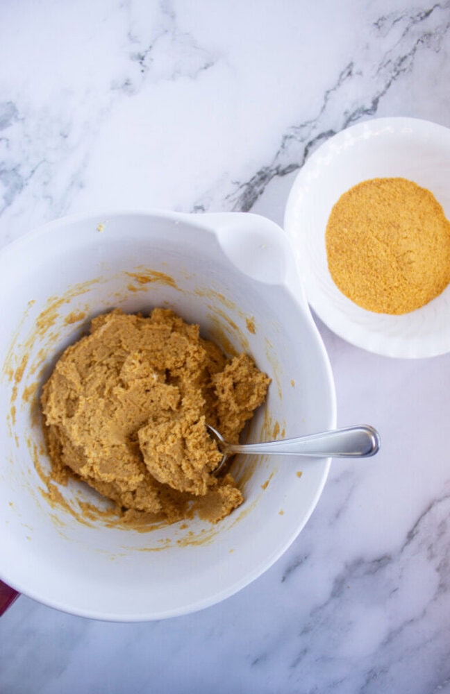 A white bowl with cookie dough and a spoon beside another bowl containing cookie crumbs on a marble surface.