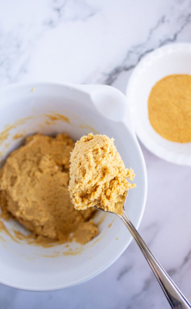 Bowl with cookie dough mixture and a spoon. A plate with brown sugar is in the background on a marble surface.