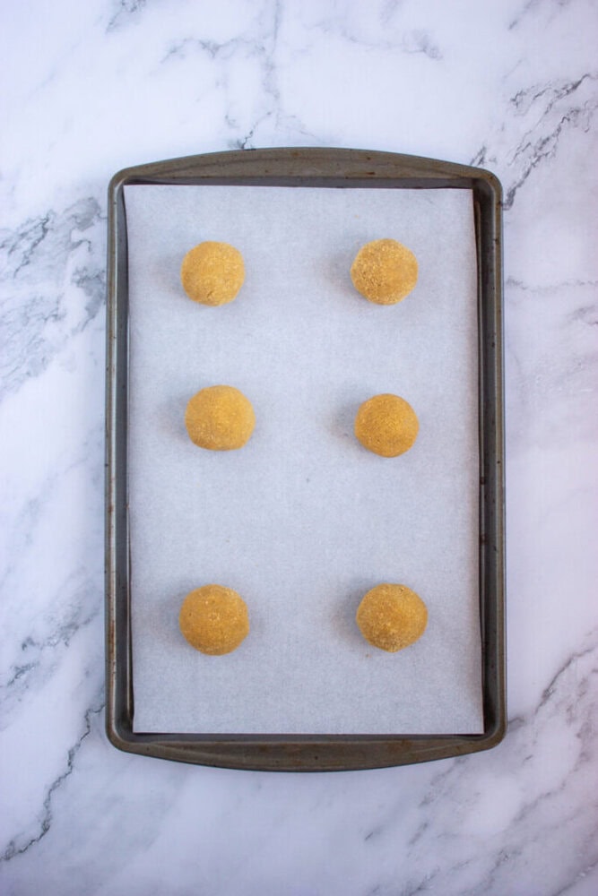 Six dough balls evenly spaced on a parchment-lined baking sheet set on a marble surface.