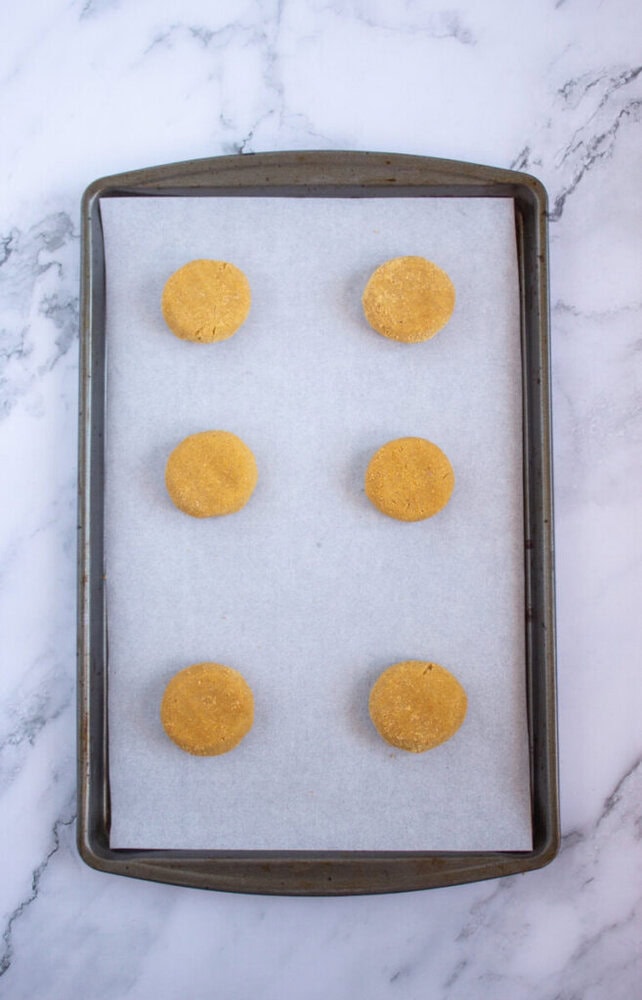 Six unbaked cookies evenly spaced on a parchment-lined baking sheet. The background is a marble surface.