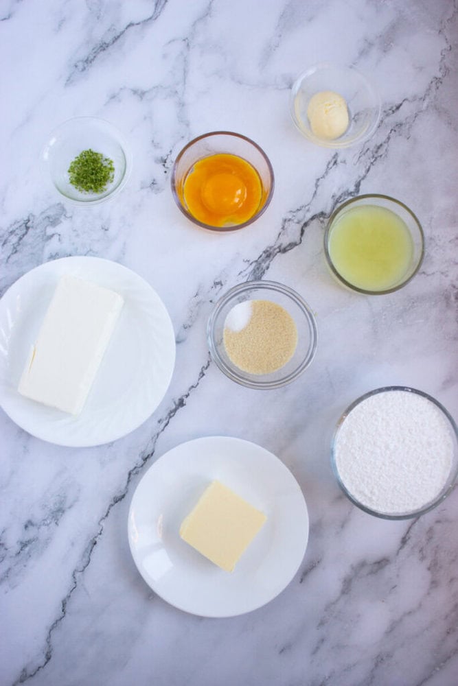 Ingredients on a marble surface include cream cheese, butter, sugar, lime zest, egg yolk, and lime juice in separate bowls and plates.
