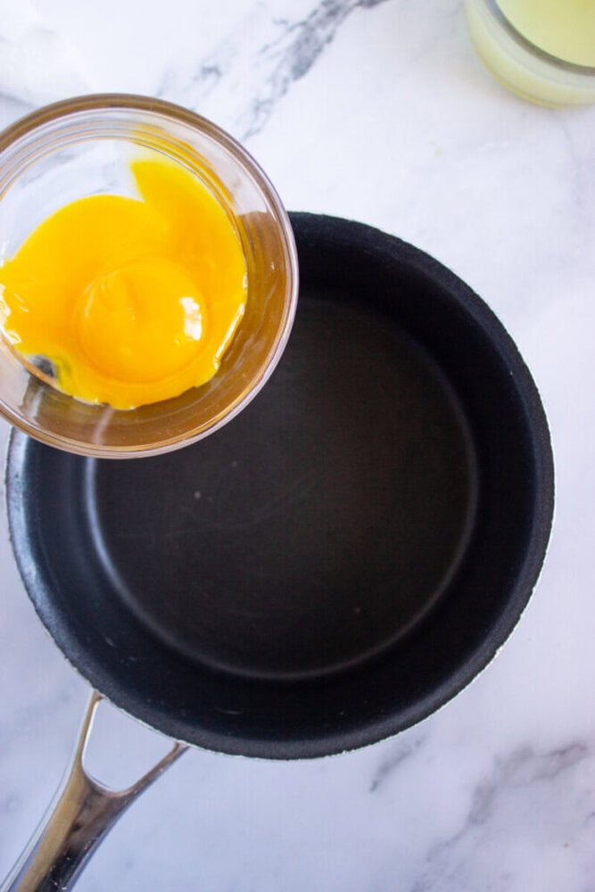 A glass bowl with egg yolks is held above an empty black saucepan on a marble surface.