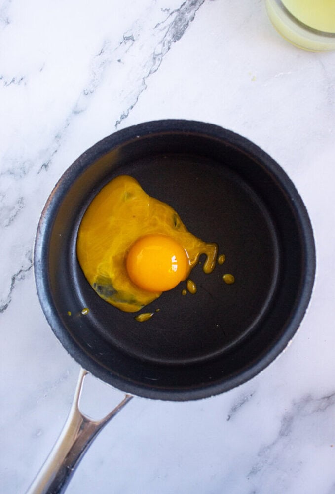 A raw egg with a broken yolk is in a small black frying pan on a marble countertop.